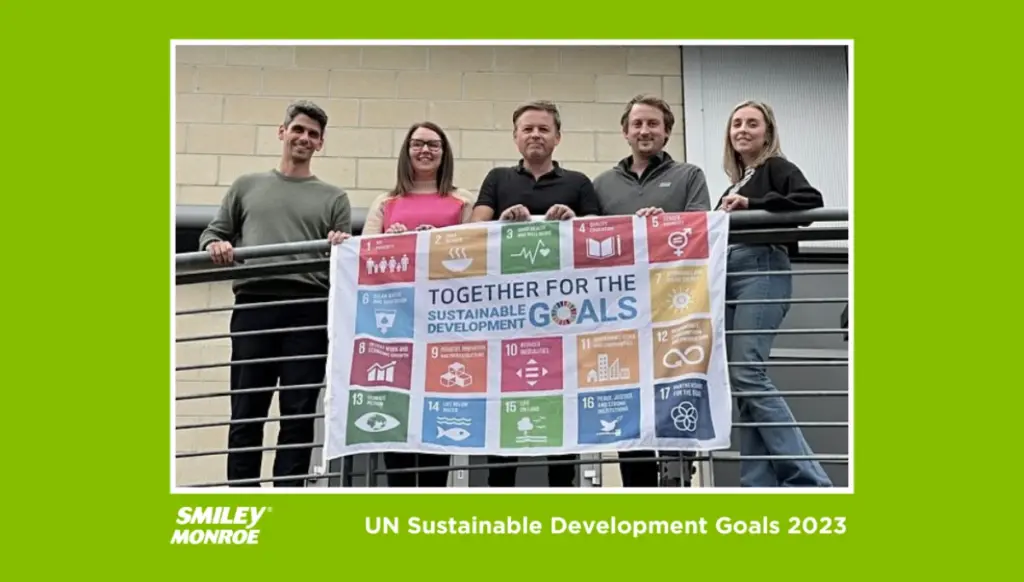 Five people standing on a balcony smiling down at the camera, holding a flag displaying the United Nations Sustainable Development Goals logo and icons and text for goals 1-17.
