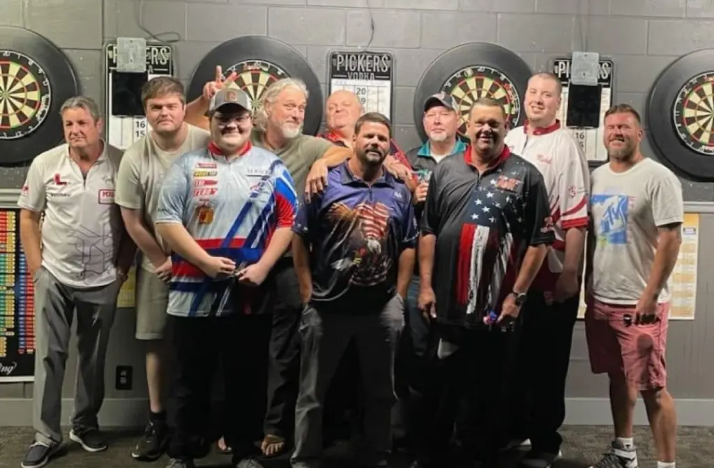 A group of men standing in front of a wall with dart boards and posters on it.