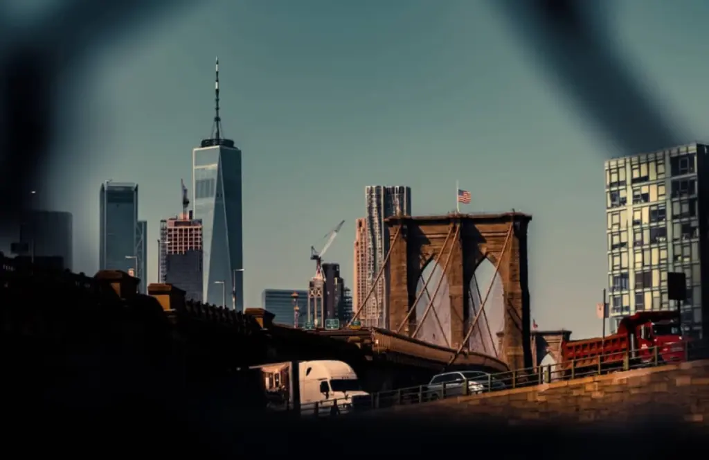 View of the Brooklyn Bridge with the Manhattan skyline in the background, including the One World Trade Center, under a clear sky, framed by an out-of-focus object in the foreground.