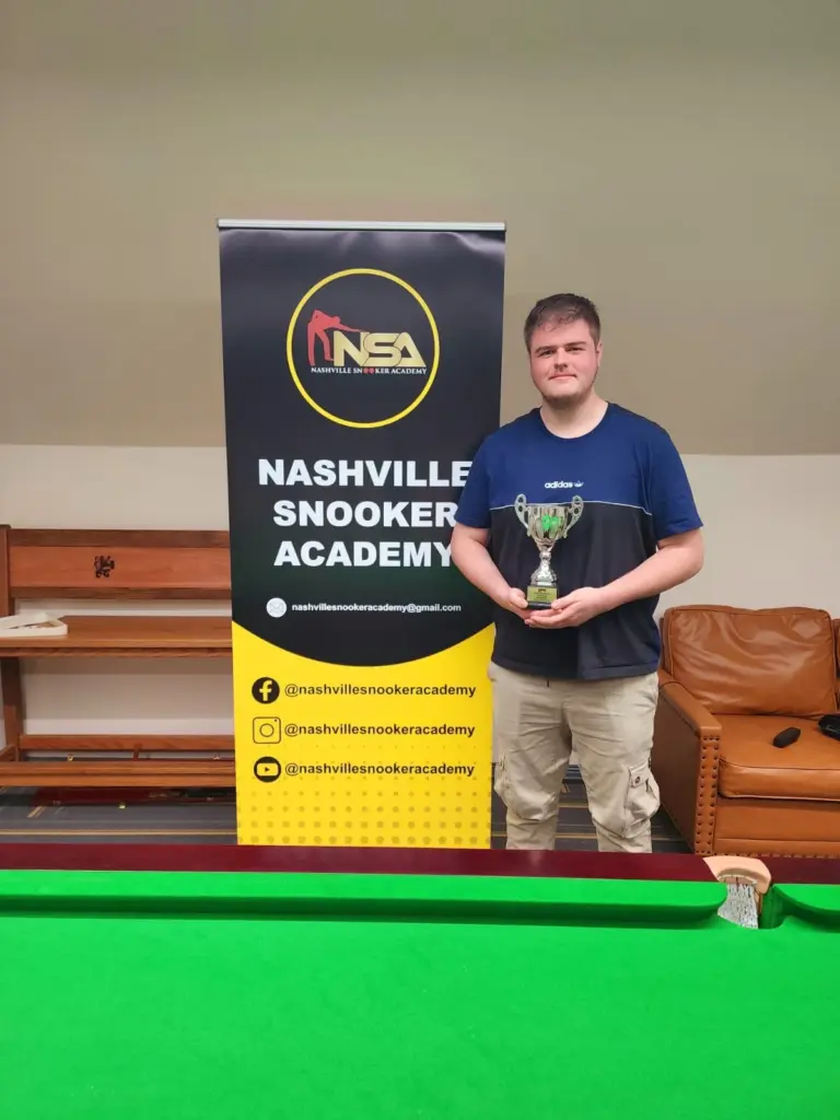 A man standing in front of a snooker table, holding a trophy beside a sign 'Nashville Snooker Academy'