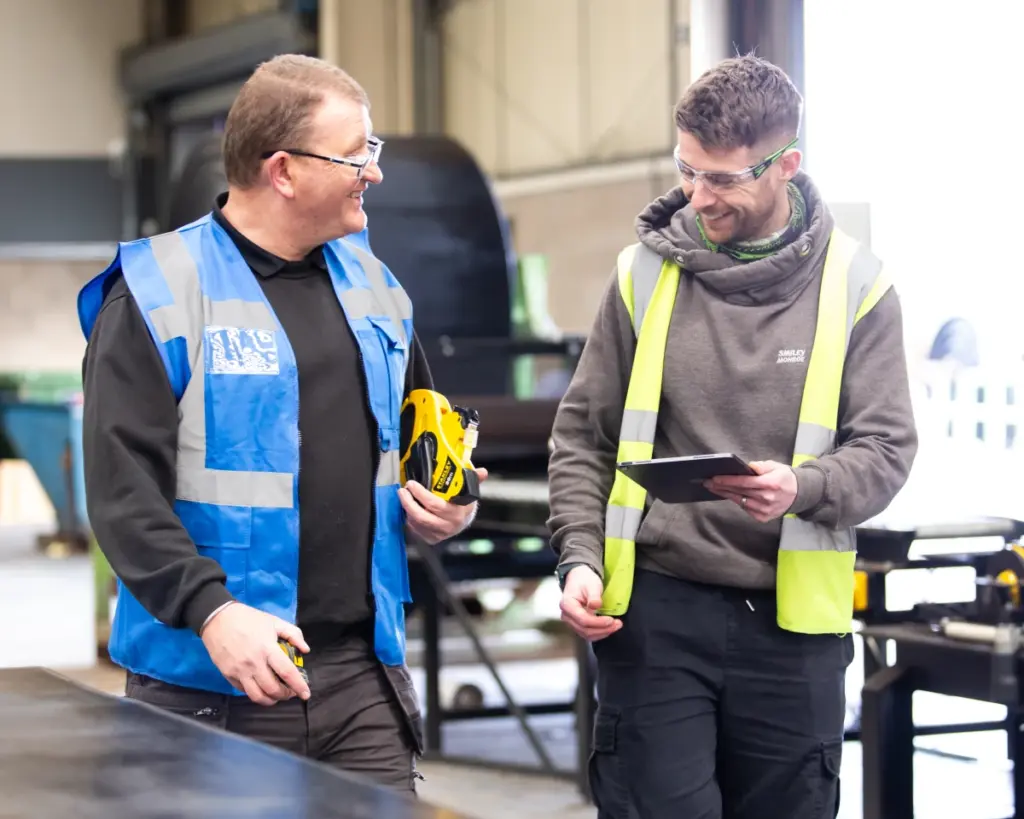 Two men wearing safety vests and goggles smile while talking in a workshop. One holds a tape measure, the other holds a tablet. Industrial equipment and tools are visible in the background.