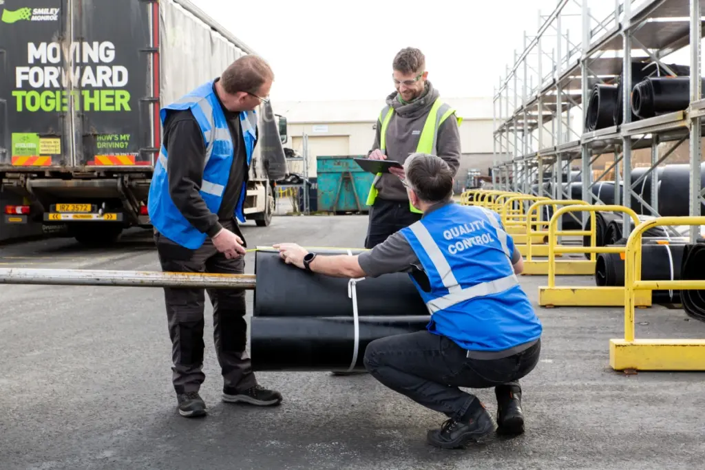 Three workers wearing blue vests labeled Quality Control inspect and discuss a large rolled material outdoors near trucks and metal racks in an industrial area. One person is holding a clipboard and taking notes.
