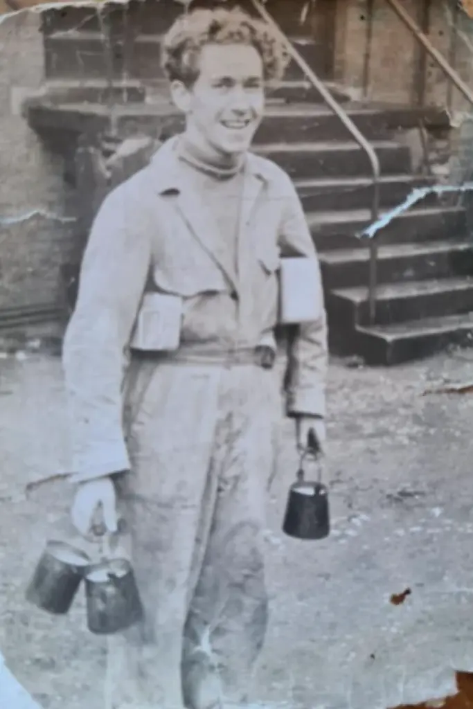 A black and white image of a man smiling at the camera wearing overalls, holding several small buckets.
