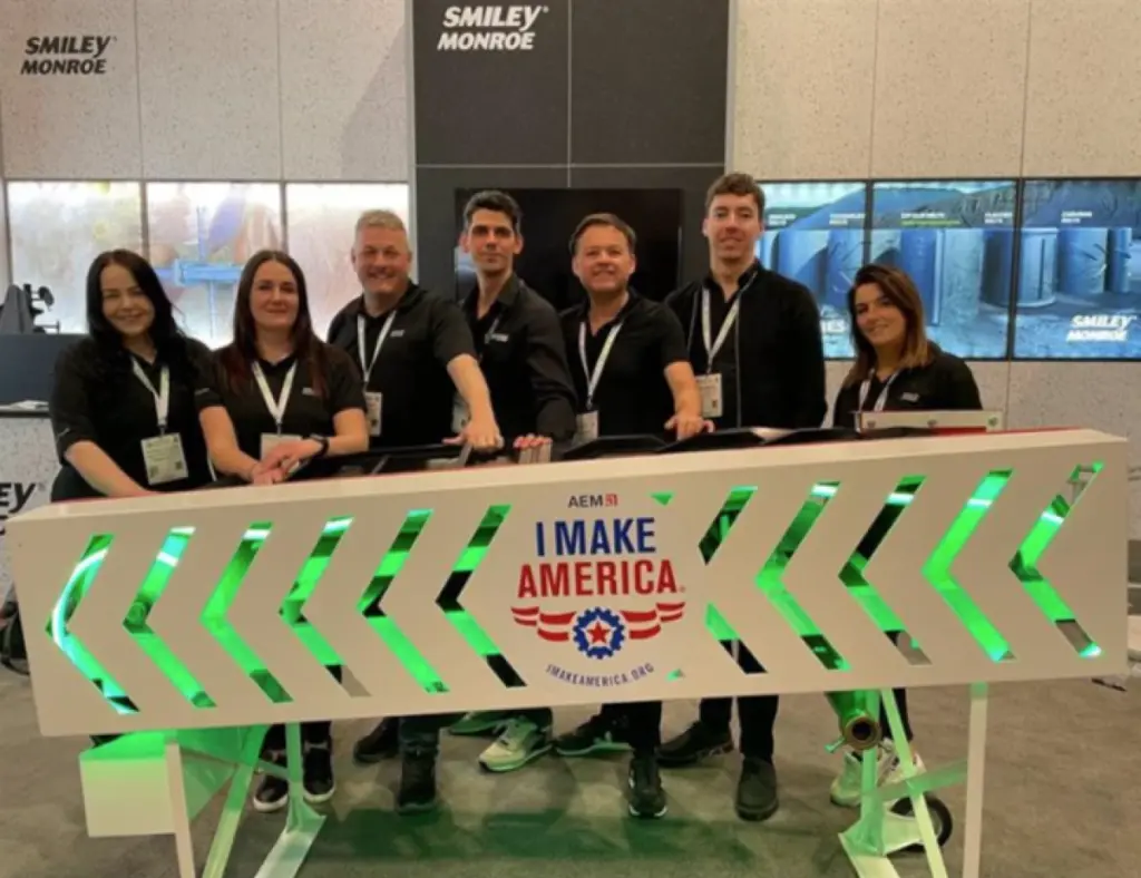 A team smiling at the camera, posing behing a board 'AEM51: I MAKE AMERICA'. The team are wearing matching black polo shirts and lanyards, and are standing in front of a wall that says Smiley Monroe.