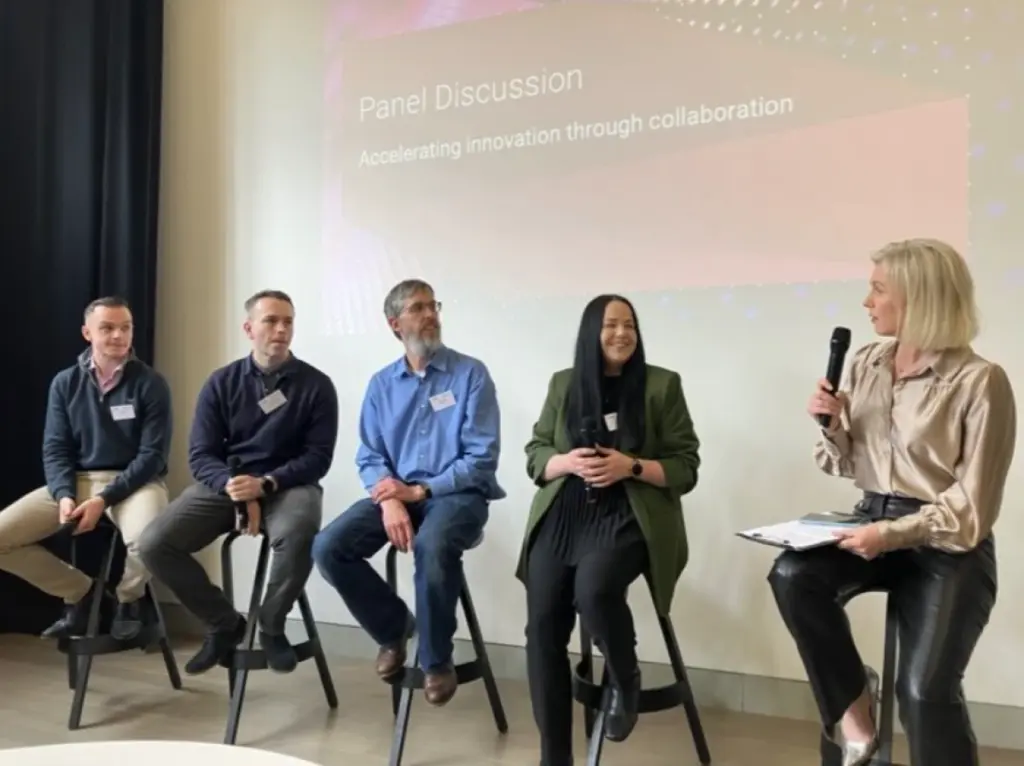 A panel discussion about 'accelerating innovation through collaboration'. A woman with a microphone and clipboard talks to a panel of four people.