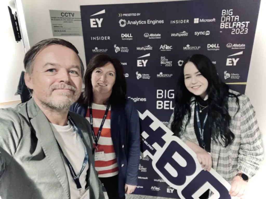 A selfie of a man and two women standing in front of a logo board at 'BIG DATA BELFAST 2023'. One woman is holding a #BD sign