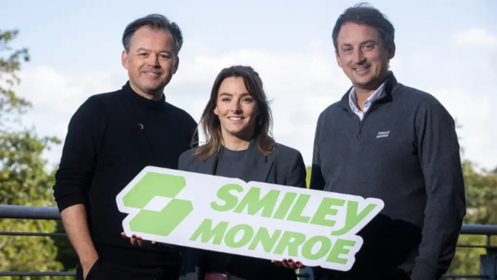 Image of three people smiling at the camera, holding a large cardboard cutout featuring SMILEY MONROE logo.
