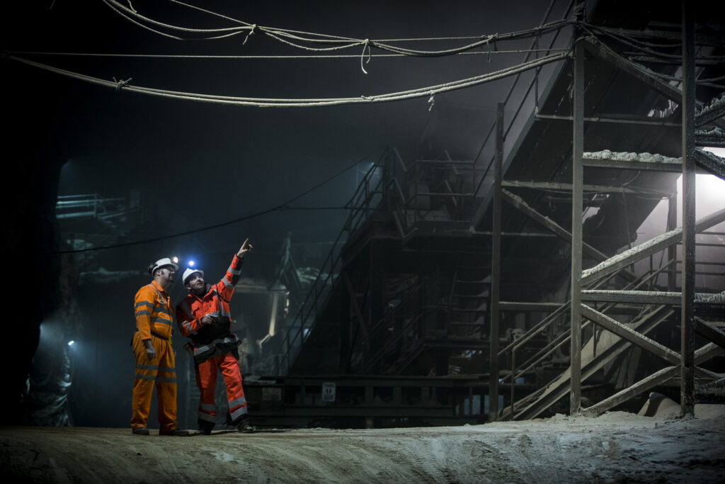 Two workers in orange safety gear and helmets stand in an industrial, dimly lit underground facility. One points upward while discussing, with large machinery and cables visible in the background.