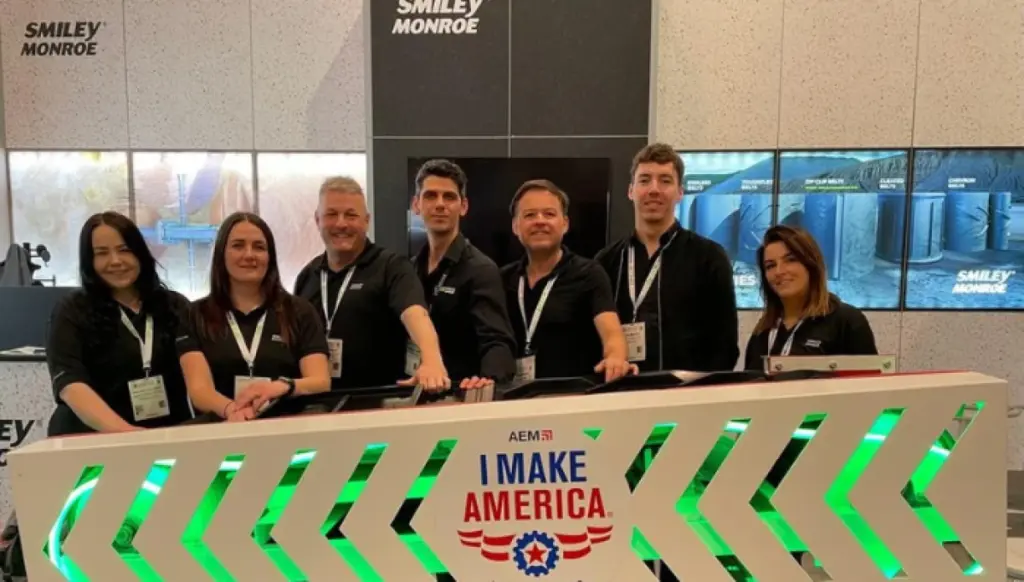 A team smiling at the camera, posing behing a board 'AEM51: I MAKE AMERICA'. The team are wearing matching black polo shirts and lanyards, and are standing in front of a wall that says Smiley Monroe.