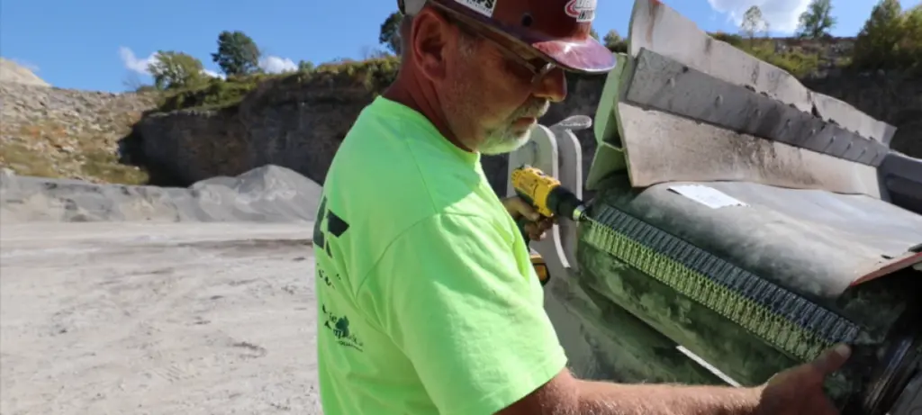 A man wearing a neon yellow shirt and hard hat uses a power drill to repair or install a component on industrial machinery at a quarry site, with piles of gravel and rock in the background.