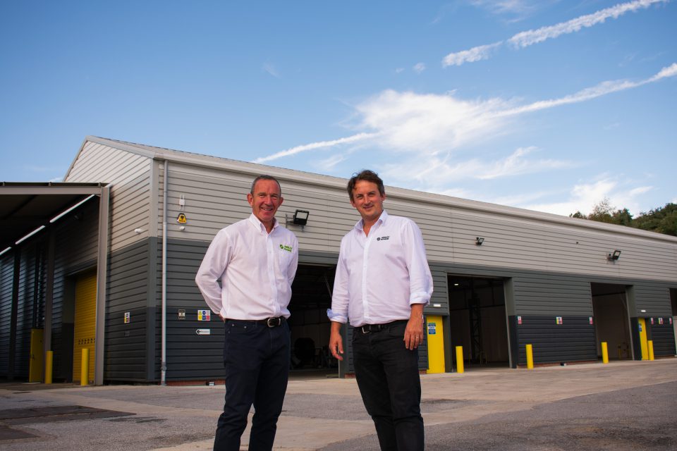 Two men in white shirts and dark trousers stand smiling outside a large industrial warehouse building on a sunny day, with blue sky and white clouds in the background.